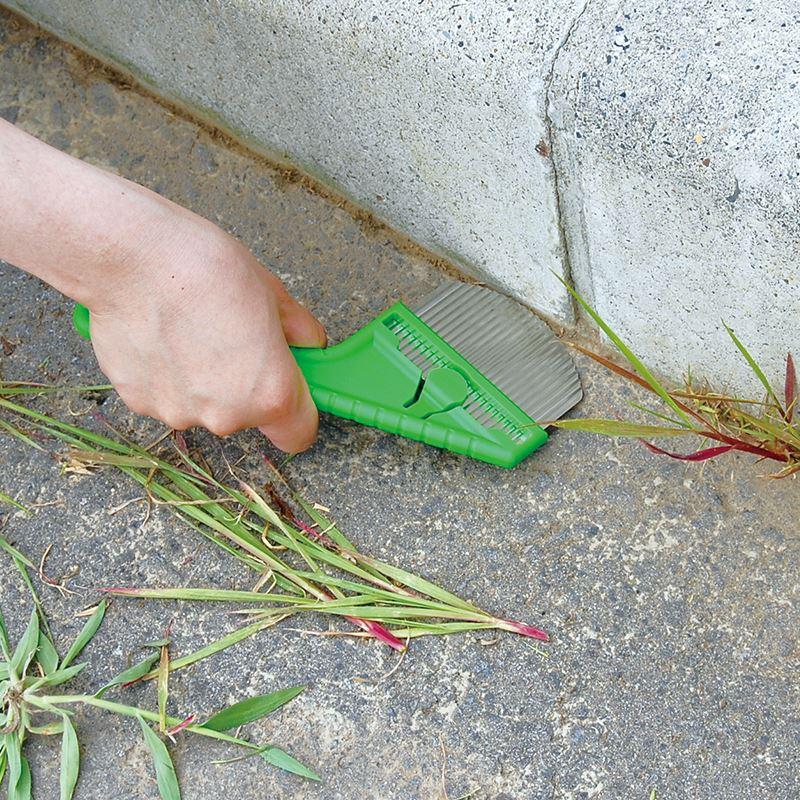 Nisaku Zassou Burush 3-inch Stainless Steel Rounded Weeder being used to get rid of weeds between the ground and a wall.